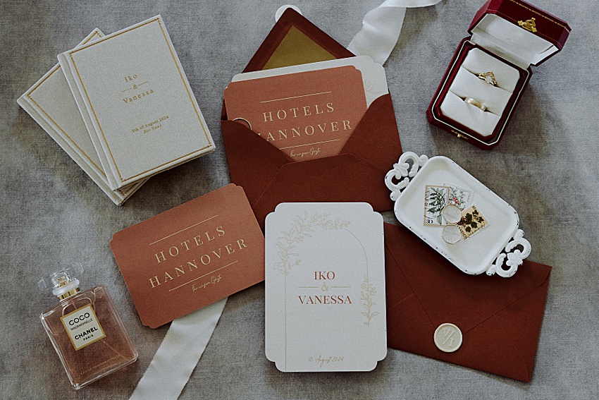 Hochzeitseinladungen und Antwortkarten auf strukturierter Oberfläche heben den goldenen Schriftzug „Iko und Vanessa“ hervor. Daneben liegen eine rote Ringschachtel, dekorative Briefmarken und ein Chanel-Parfümflakon. Dunkelrote Umschläge geben den Ton für die elegante Hochzeit in Hannover an.