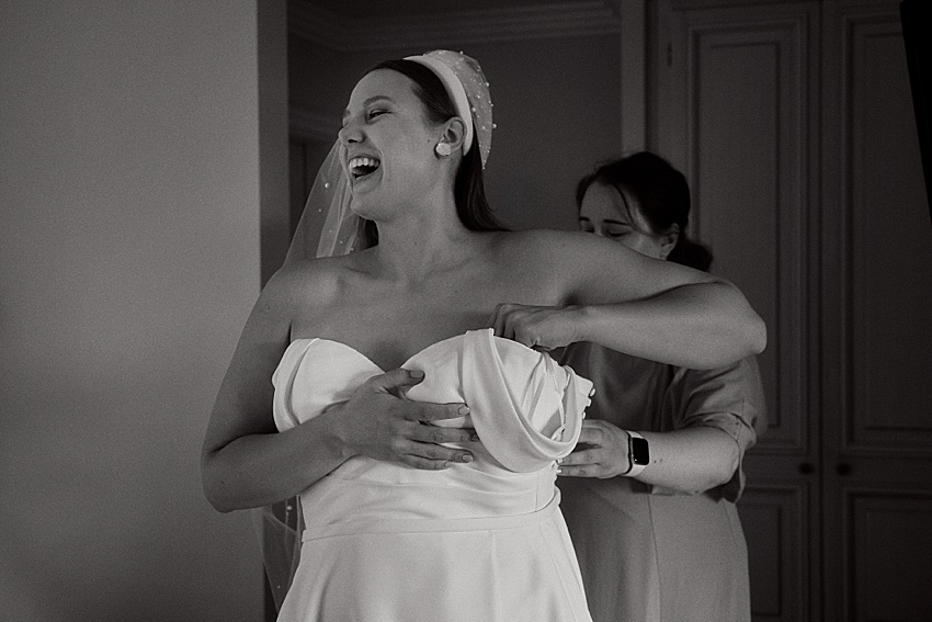 Eine Braut lächelt breit, während ihr jemand bei einer Hochzeit in Hannover hilft, ihr trägerloses Brautkleid zu richten. Sie trägt einen mit kleinen Perlen verzierten Schleier. Die Szene spielt in einem Raum, nahe einer geschlossenen Tür, und das Bild fängt die zeitlose Eleganz in Schwarzweiß ein.