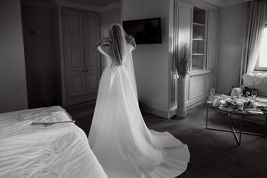 Eine Braut im Brautkleid steht mit abgewandtem Blick in einem Hotelzimmer im Hotel Kaiserhof. Das Zimmer verfügt über ein gemachtes Bett, einen an der Wand montierten Fernseher und einen Tisch mit leeren Gläsern und Geschirr. Sonnenlicht fällt durch das Fenster rechts. Das Bild ist in Schwarzweiß gehalten und vermittelt zeitlose Eleganz.