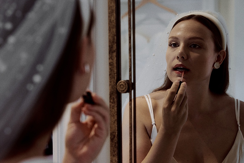 Eine Frau in weißem Kleid und Haarband trägt Lippenstift auf und blickt dabei in den Spiegel – eine Erinnerung an eine Braut, die sich auf ihre Hochzeit vorbereitet. Ihr Spiegelbild spiegelt Eleganz vor dem weichen Hintergrund der Kleiderbügel wider und erinnert an den zeitlosen Charme von Orten wie den Herrenhäuser Gärten.
