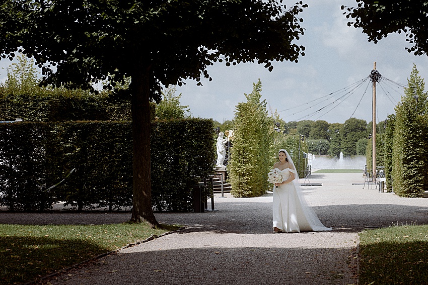 Eine Braut in weißem Kleid und Schleier schlendert mit einem Blumenstrauß in der Hand durch die bezaubernden Herrenhäuser Gärten. Gepflegte Hecken, majestätische Bäume und ein Brunnen zieren die Landschaft unter Hannovers leicht bewölktem Himmel, als hätte das Hotel Kaiserhof auf ihren Empfang gewartet.