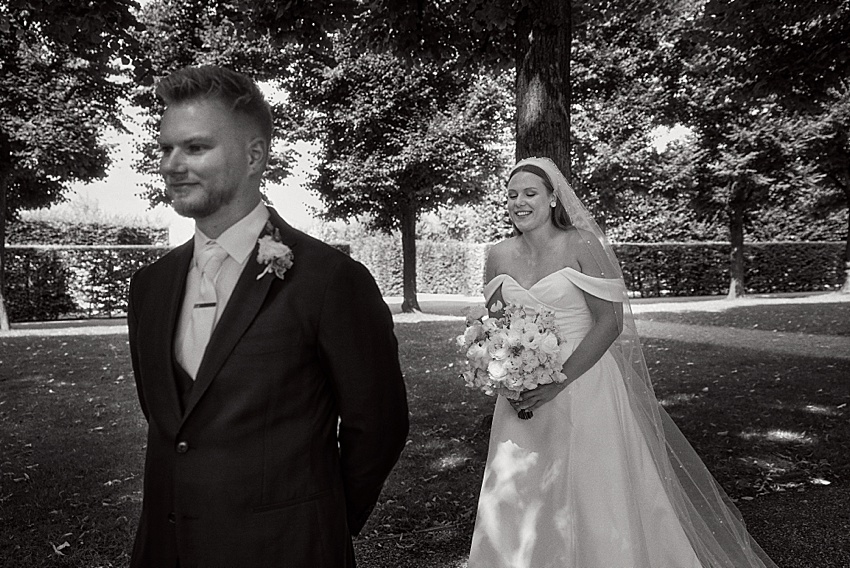 Eine Braut mit Blumenstrauß in der Hand und schulterfreiem Brautkleid lächelt, als sie sich ihrem Bräutigam im Anzug in den Herrenhäuser Gärten nähert. Umgeben von Bäumen und Sonnenlicht fühlt sich ihre Hochzeit in Hannover wie ein Traum an. Der Bräutigam steht mit dem Rücken zur Braut, und die Vorfreude steigt in der ruhigen Umgebung.