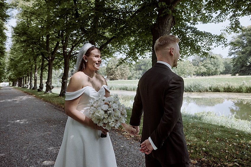 Braut und Bräutigam stehen unter einer Baumreihe neben einem Teich in den üppigen Herrenhäuser Gärten in Hannover. Die Braut in einem schulterfreien weißen Kleid lächelt und hält einen Brautstrauß in der Hand. Der Bräutigam, in einem dunklen Anzug, blickt abgewandt und hat die Hände hinter dem Rücken verschränkt.