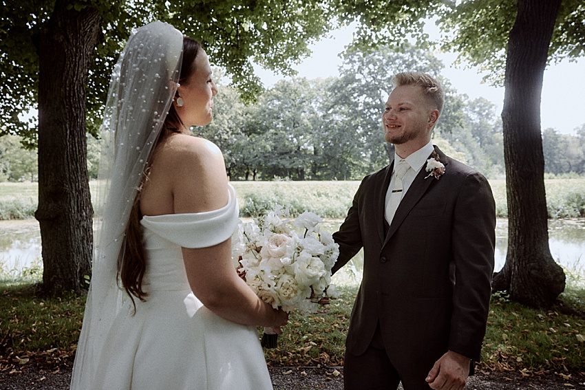 Im Schatten der Bäume in den Herrenhäuser Gärten hält eine Braut in einem schulterfreien weißen Kleid einen Blumenstrauß in den Händen. Der Bräutigam, elegant gekleidet in einem braunen Anzug, blickt sie lächelnd an. Diese bezaubernde Szene ist Teil einer traumhaften Hochzeit in Hannover.