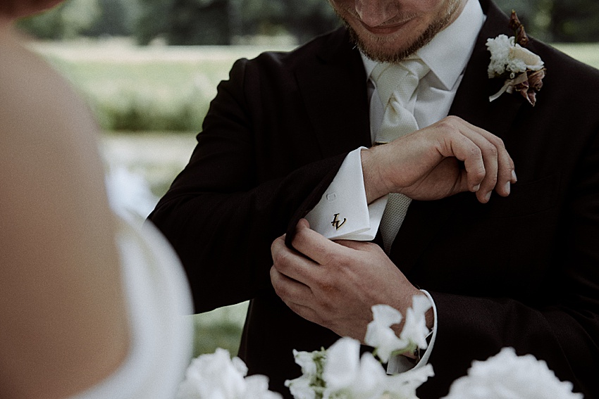 Ein Mann im dunklen Anzug rückt seinen mit Initialen gravierten Manschettenknopf zurecht. Elegant gekleidet für eine Hochzeit in Hannover. Er trägt eine weiße Krawatte und eine Ansteckblume und steht inmitten üppigen Grüns, das an die Herrenhäuser Gärten erinnert. Neben ihm vervollständigt eine weitere Figur in einem weißen Kleid die malerische Szene.