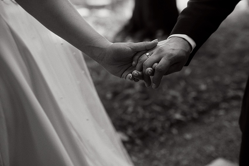 Ein Schwarz-Weiß-Bild zeigt zwei Personen, die sich bei ihrer Hochzeit in Hannover Händchen halten. Die eine trägt ein Kleid, die andere einen Anzug. Der Fokus liegt auf ihren Händen und einem sichtbaren Ehering. Der sanft unscharfe Hintergrund erinnert an die Eleganz der Herrenhäuser Gärten.