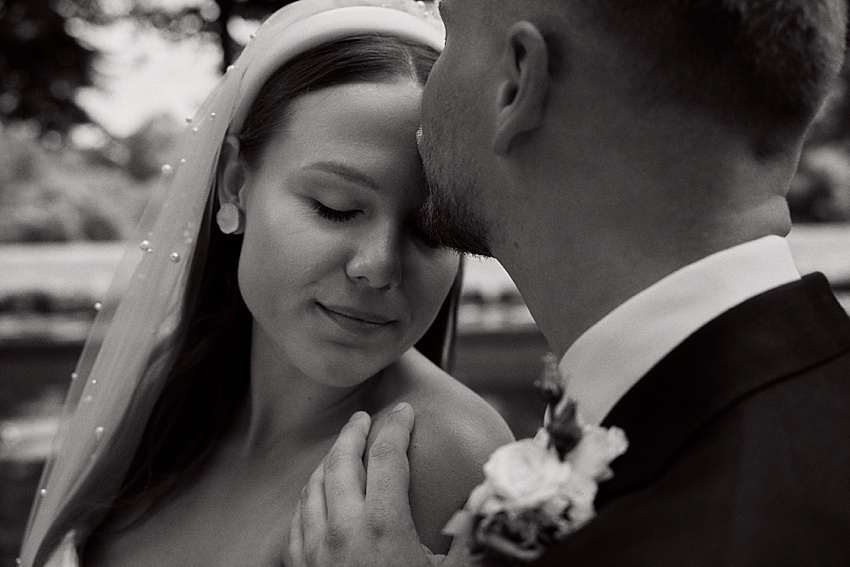 Ein Schwarz-Weiß-Foto zeigt ein Paar, das sich während seiner Hochzeit in Hannover in den malerischen Herrenhäuser Gärten umarmt. Die Frau trägt einen Brautschleier und Ohrringe, während der Mann, der einen Anzug mit Ansteckblume trägt, sanft ihre Schulter hält, während sie ihre Köpfe aneinander lehnen.