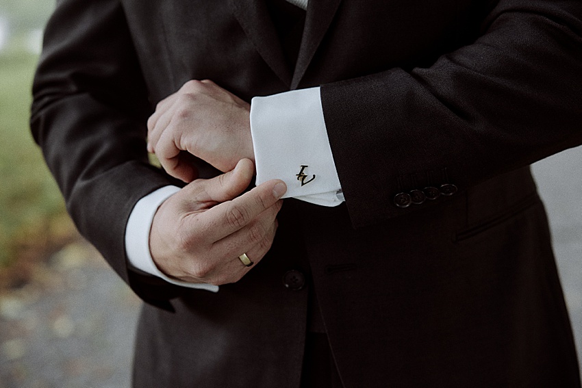 In einem eleganten dunklen Anzug rückt eine Person einen weißen Manschettenknopf mit dem stilisierten Buchstaben „Z“ am Ärmel zurecht. An der linken Hand glänzt ein goldener Ehering, der auf eine kürzlich erfolgte Hochzeit hindeutet. Der unscharfe Hintergrund erinnert an die Ruhe der Herrenhäuser Gärten in Hannover.