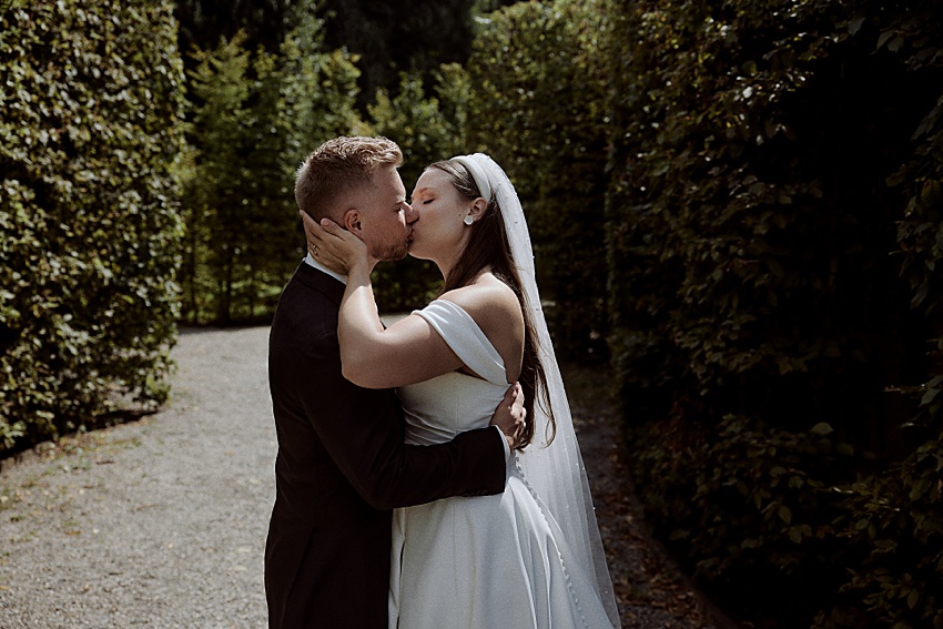 Ein Paar im Hochzeitskleid küsst sich draußen in den Herrenhäuser Gärten. Die Braut in weißem Kleid und Schleier steht neben dem Bräutigam im dunklen Anzug. Sie sind umgeben von hohen, grünen Hecken entlang eines Kieswegs und fangen die Atmosphäre einer romantischen Hochzeit in Hannover ein.