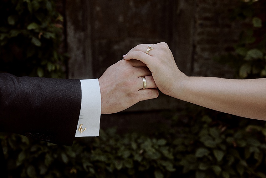 Eine Nahaufnahme zeigt zwei gefaltete Hände bei einer Hochzeit in Hannover. Die linke Hand, geschmückt mit monogrammierten Manschettenknöpfen, gehört jemandem im schwarzen Anzug, während die rechte Hand einen goldenen Ring mit einem kleinen Diamanten trägt. Im Hintergrund erinnert üppiges grünes Laub an die ruhige Schönheit der Herrenhäuser Gärten.