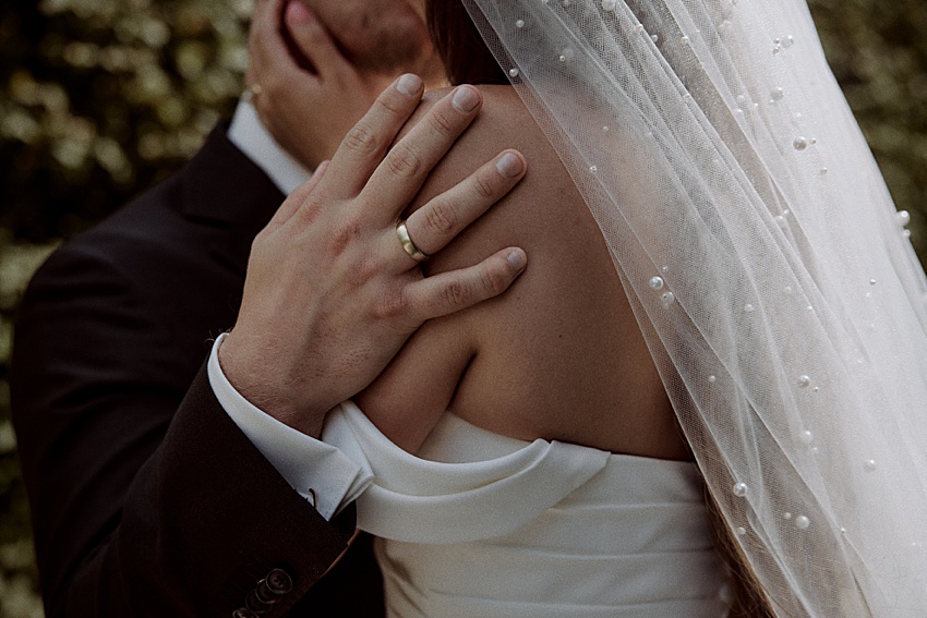 Inmitten der ruhigen grünen Herrenhäuser Gärten umarmt sich ein Paar in Hochzeitskleidung. Die eine, in dunklem Anzug und mit Ehering, hält die andere am Hals. Die andere, in einem weißen Kleid mit perlenbesetztem Schleier, der ihr den Rücken zukehrt, rundet diesen bezaubernden Hochzeitsmoment in Hannover ab.