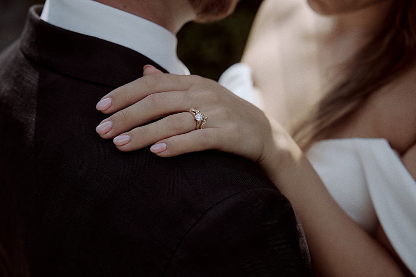 Eine Frau mit langen Haaren und einem trägerlosen weißen Outfit legt ihre Hand auf die Schulter eines Mannes im dunklen Anzug. Ihr goldener Ring glänzt dezent, während die beiden ihre Hochzeit in Hannover feiern. Das Bild fängt die Eleganz des Augenblicks ein und hebt Schultern und Hände vor dem romantischen Hintergrund hervor.