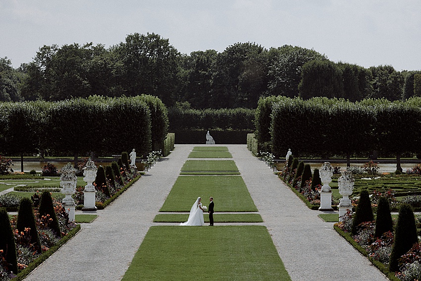 Braut und Bräutigam stehen auf einem breiten, grasbewachsenen Weg in einem formalen Garten der Herrenhäuser Gärten. Gepflegte Hecken und Bäume säumen den Weg. Steinstatuen und dekorative Pflanzen stehen zu beiden Seiten, mit einem Waldgebiet im Hintergrund – die perfekte Kulisse für eine Hochzeit in Hannover.