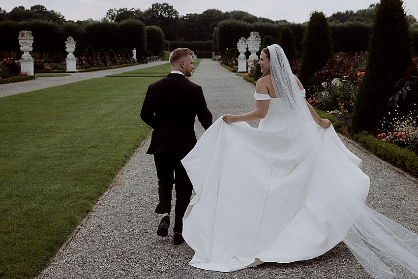 Braut und Bräutigam spazieren einen Kiesweg in den Herrenhäuser Gärten entlang. Die Braut, in einem weißen schulterfreien Kleid mit langem Schleier, und der Bräutigam, im schwarzen Anzug, genießen ihre Hochzeit in Hannover. Grüne Rasenflächen und gepflegte Gärten mit Statuen säumen ihren malerischen Weg.