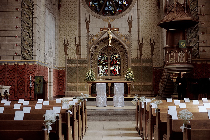 Innenraum einer geschmückten Kirche in Hannover mit einem reich verzierten Altar mit Blumenarrangements und einem Kruzifix. Reihen von Holzbänken mit weißen Schleifen an den Gangplätzen führen dorthin. Buntglasfenster und detailreiche Schnitzereien schmücken die Wände und erinnern an eine Hochzeit in den Herrenhäuser Gärten.