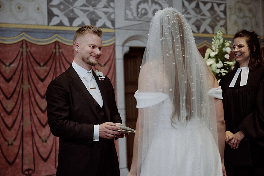 Bei einer Hochzeit in Hannover stehen sich Braut und Bräutigam inmitten der kunstvoll dekorierten Kulisse gegenüber. Der Bräutigam im dunklen Anzug hält eine Karte, während seine Braut in ihrem schulterfreien, perlenbesetzten Kleid glänzt. Daneben rundet ein lächelnder Standesbeamter die bezaubernde Szene in den Herrenhäuser Gärten ab.
