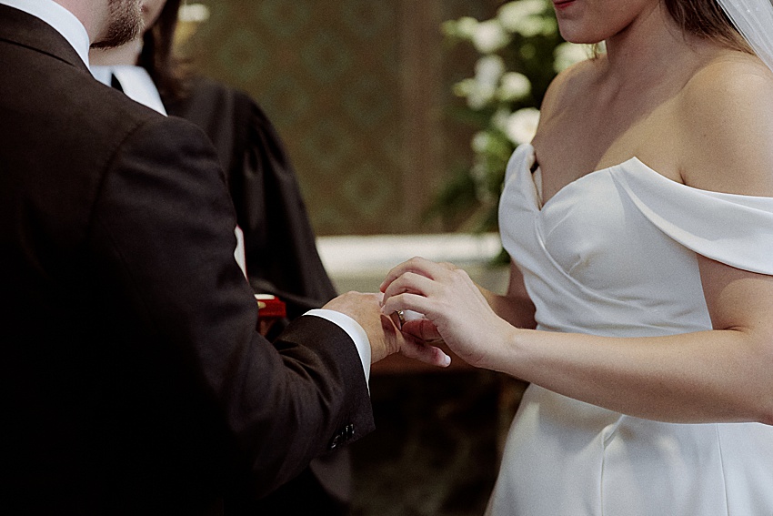 Bei einer Hochzeit in Hannover tauschen Braut und Bräutigam die Ringe. Die Braut glänzt in ihrem weißen, schulterfreien Kleid, während der Bräutigam ihre Eleganz in einem eleganten schwarzen Anzug unterstreicht. In der Nähe, unter dem Blick eines Standesbeamten in schwarzer Robe, akzentuieren weiße Blumen aus den Herrenhäuser Gärten die Szene.