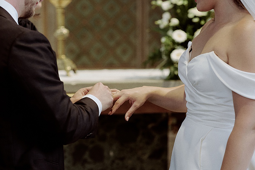 Bei einer Hochzeit in Hannover steckt eine Person im schwarzen Anzug einer anderen Person einen Ring an den Finger, während sie ein weißes, schulterfreies Brautkleid trägt. Sie stehen vor einem mit weißen Blumen geschmückten Altar. Die Romantik dieses Augenblicks wird nur durch die Eleganz der Herrenhäuser Gärten übertroffen.