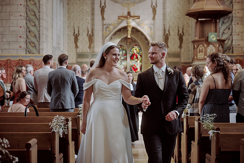 Braut und Bräutigam schreiten Hand in Hand durch den Mittelgang einer Kirche, während sie von ihren Flitterwochen im Hotel Kaiserhof träumen. Das schulterfreie weiße Kleid der Braut harmoniert perfekt mit dem eleganten Anzug des Bräutigams, während die Gäste in den Holzbänken sie unter dem sanften Blick eines Kreuzes bewundern.