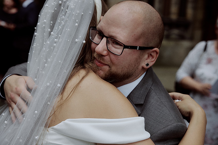 Eine Braut in einem weißen Kleid mit Perlenverzierungen umarmt einen glatzköpfigen Mann mit Brille und grauem Anzug bei ihrer Hochzeit in Hannover. Vor der lebendigen Kulisse der Herrenhäuser Gärten verleiht die verschwommene Menschenmenge diesem besonderen Moment eine festliche Atmosphäre.