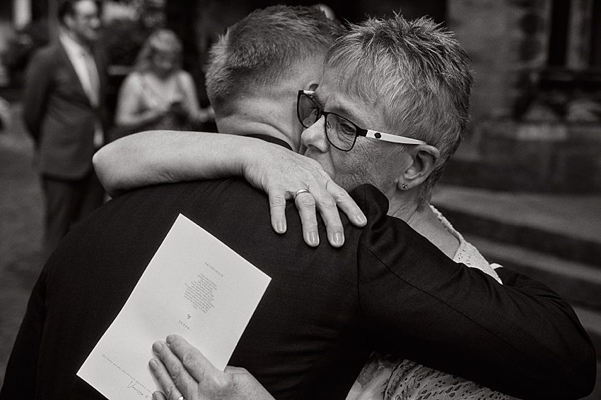 Ein Schwarz-Weiß-Foto zeigt eine enge Umarmung zwischen einem Mann im Anzug und einer älteren Frau mit Brille bei Hochzeit in Hannover. Der Mann hält einen Zettel mit aufgedrucktem Text, möglicherweise vom Hotel Kaiserhof. Weitere Personen in formeller Kleidung sind im Hintergrund unscharf zu sehen.