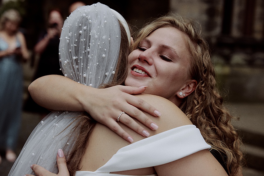 Zwei Frauen umarmen sich bei einer Hochzeit im Freien in den wunderschönen Herrenhäuser Gärten in Hannover. Die Frau, die der Kamera zugewandt ist, hat langes, lockiges Haar und lächelt mit geschlossenen Augen. Die Frau, die der Kamera den Rücken zuwendet, trägt ein weißes Gewand und einen perlenbesetzten Schleier, während die anderen im Hintergrund verschwimmen.