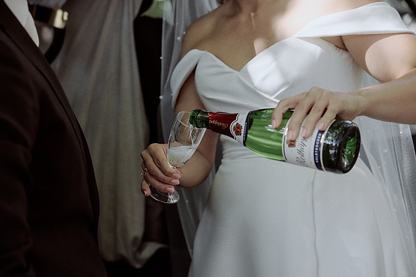 Bei einer Hochzeit in Hannover schenkt eine Braut im weißen, schulterfreien Brautkleid elegant Champagner in ein Glas ein, das von einem Mann im dunklen Anzug gehalten wird. Die Flasche mit grünem Etikett und roten Akzenten lässt auf eine intime Feier im Hotel Kaiserhof schließen.