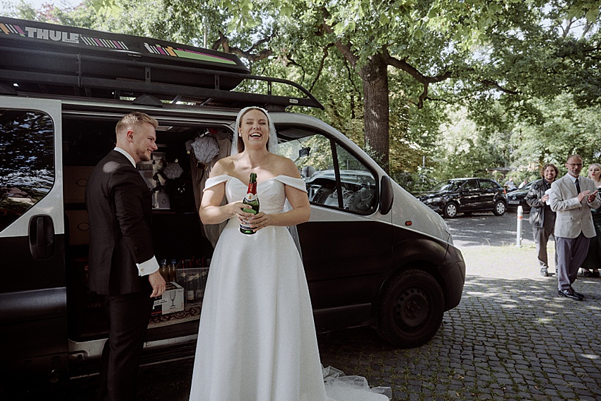 Braut und Bräutigam stehen neben einem schwarzen Van mit Dachgepäckträger und bereiten sich auf ihre Hochzeit in Hannover vor. Die Braut in ihrem weißen schulterfreien Kleid hält eine Flasche in der Hand und lacht, während der Bräutigam in seinem eleganten schwarzen Anzug neben ihr steht. Im Hintergrund sind parkende Autos und Menschen im Schatten der Bäume zu sehen.