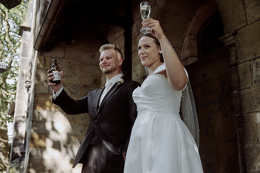 Ein Bräutigam im dunklen Anzug hält eine Bierflasche, während eine Braut im weißen Kleid mit ihrem Champagnerglas anstößt. Sie stehen vor einem Steingebäude mit Holztüren, umgeben von grünen Bäumen, die den Charme der Herrenhäuser Gärten widerspiegeln.