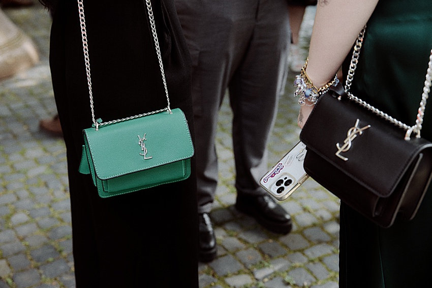 Zwei Personen stehen mit Saint Laurent-Kettentaschen, eine grüne und eine schwarze, jeweils mit dem ikonischen YSL-Logo. Inmitten des Kopfsteinpflasters vor dem eleganten Hotel Kaiserhof hält eine von ihnen ein Smartphone mit farbenfrohem Design – ein perfekter Stilmix für jede Hochzeit in Hannover.