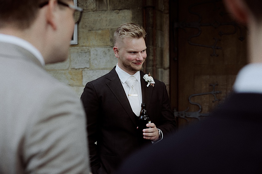 Ein Mann im Anzug steht mit einer Bierflasche da und lächelt leicht. Mit seinen kurzen blonden Haaren und einer Ansteckblume am Revers strahlt er Charme aus. Die Kulisse erinnert an Hannovers elegante Herrenhäuser Gärten, mit Andeutungen zweier weiterer Personen im Vordergrund nahe der Steinmauer und der Holztür.
