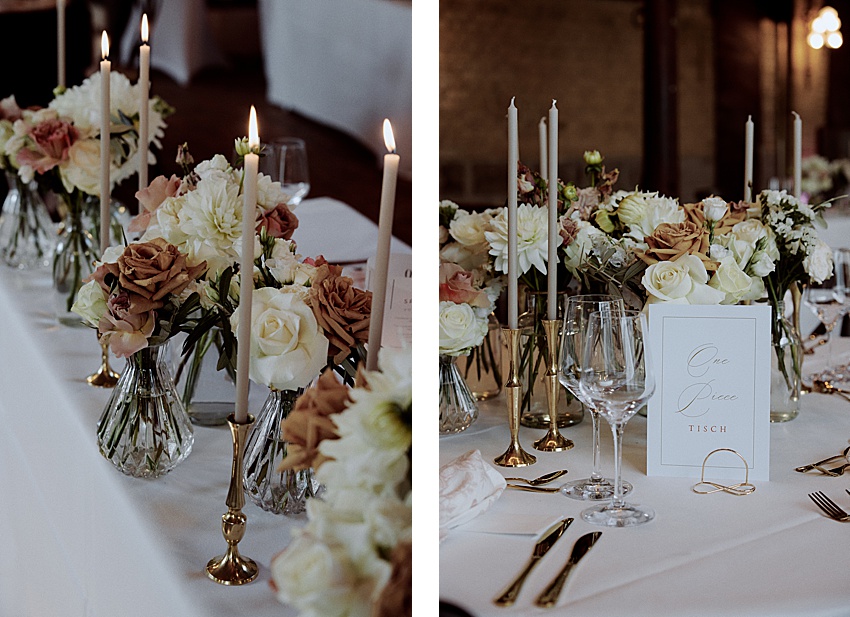 Ein gedeckter Tisch mit weißer Tischdecke präsentiert brennende Stabkerzen in goldenen Haltern, Blumenarrangements mit weißen und rosa Rosen und eine Karte mit der Aufschrift „One Piece Tisch“. Weingläser und Besteck sind ordentlich arrangiert, ganz im Stil einer eleganten Hochzeit im Hotel Kaiserhof.
