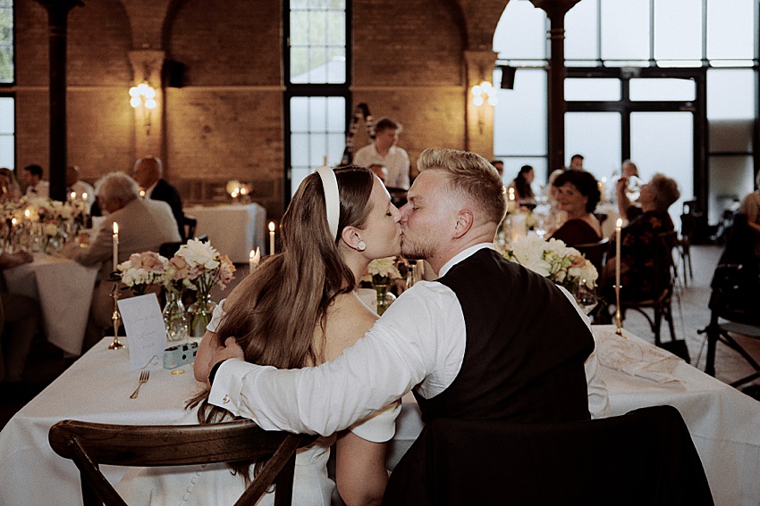 Bei einer Hochzeit in Hannover küsst sich ein Paar zärtlich an seinem elegant gedeckten Tisch, umgeben von Gästen und dem Zauber von Blumen und Kerzen. Die großen Fenster und Backsteinwände des Raumes tragen zum gehobenen Ambiente bei.