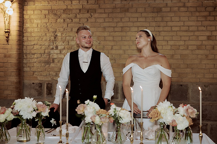 Ein Paar steht hinter einem mit Kerzen und Blumenvasen geschmückten Tisch und verkörpert die Romantik einer Hochzeit in Hannover. Der Mann trägt eine schwarze Weste über einem weißen Hemd, während die Frau ein schulterfreies weißes Kleid trägt, das sich vom charmanten Ambiente der Backsteinmauer abhebt.