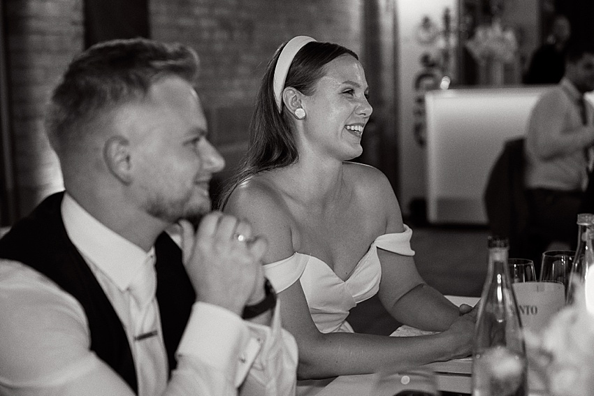 Ein Schwarz-Weiß-Foto zeigt ein lächelndes Paar bei einer Hochzeit in Hannover. Die Frau im schulterfreien Kleid und mit Haarband sitzt neben dem Mann im Anzug. Flaschen und ein Schild mit der Aufschrift „VINO“ schmücken den Tisch und lassen einen eleganten Abend erahnen, der vielleicht an die Herrenhäuser Gärten erinnert.