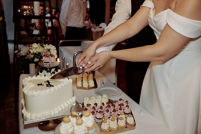 Das Paar schneidet bei seiner Hochzeitsfeier in Hannover eine weiße, rechteckige Torte an. Die Torte, umgeben von Gebäck und Blumenschmuck, steht wunderschön auf dem Tisch. Im Hintergrund beobachten die Gäste gespannt, wie die Frau in einem schulterfreien weißen Kleid die bezaubernde Atmosphäre des Hotels Kaiserhof genießt.