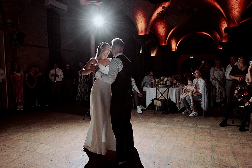 Bei einer Hochzeit im Herzen Hannovers wirbelt ein Paar anmutig im Scheinwerferlicht der Herrenhäuser Gärten. Das weiße Kleid der Braut schimmert, während ihr Bräutigam, in Weste und Krawatte gekleidet, sie unter der warm beleuchteten Gewölbedecke zwischen den Tischen der Gäste hindurchführt.