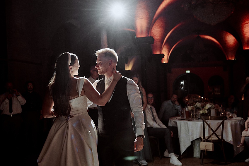 Bei der Hochzeit in Hannover tanzen Braut und Bräutigam unter den gewölbten Decken des Hotels Kaiserhof. Das weiße Kleid der Braut erstrahlt im warmen Licht, während die Gäste von blumengeschmückten Tischen aus diese zauberhafte Feier bewundern.