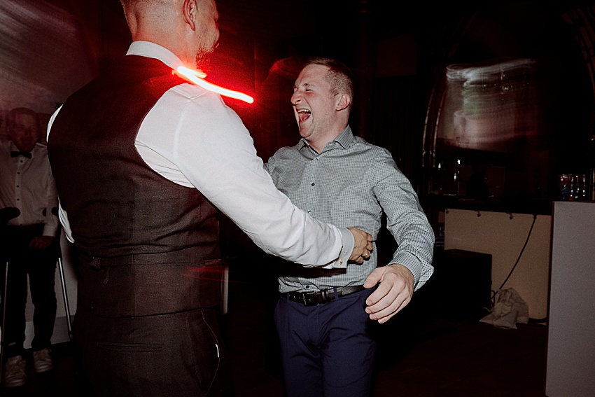Zwei Männer tanzen fröhlich bei einer Indoor-Veranstaltung, vermutlich einer Hochzeit in Hannover. Einer trägt ein weißes Hemd und eine dunkle Weste, der andere ein hell gemustertes Hemd und eine dunkle Hose. Rote Lichteffekte verstärken die ausgelassene Atmosphäre und erinnern an Feiern in den Herrenhäuser Gärten.