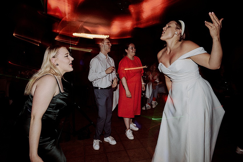 Eine Gruppe von vier Personen tanzt auf einer Hochzeit in den Herrenhäuser Gärten in Hannover. Eine Person in einem weißen Kleid singt leidenschaftlich, eine andere in einem schwarzen Kleid stimmt mit ein. Im Hintergrund stehen zwei Personen, eine in Rot, die andere in Freizeitkleidung.